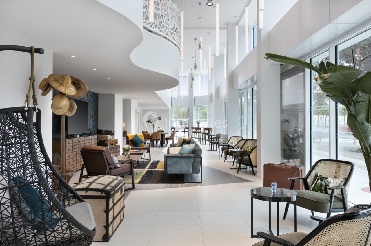 Espace intérieur lumineux avec mobilier moderne, fauteuils variés, plantes et grandes fenêtres laissant entrer la lumière…