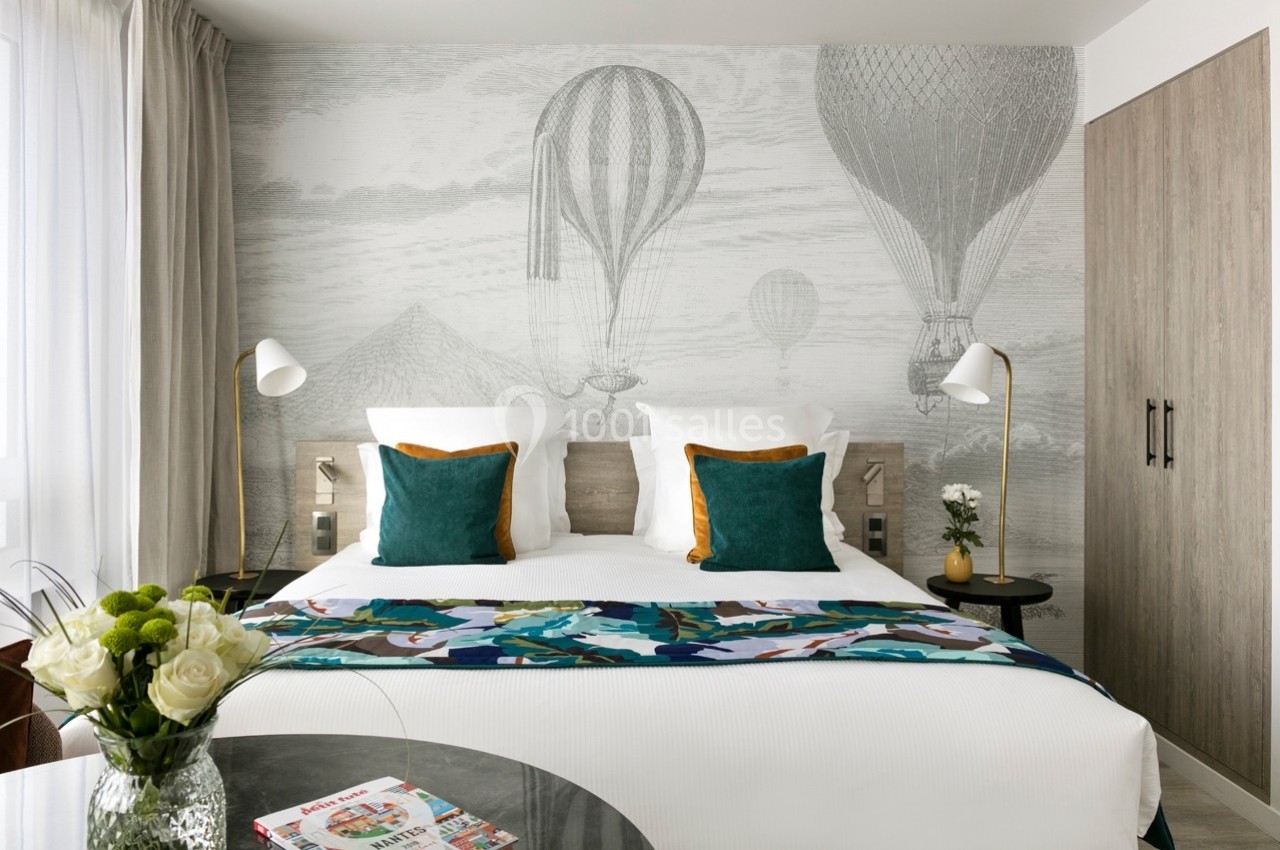 Lit double avec coussins verts, mur décoré de montgolfières dessinées, table avec fleurs et magazine au premier plan.