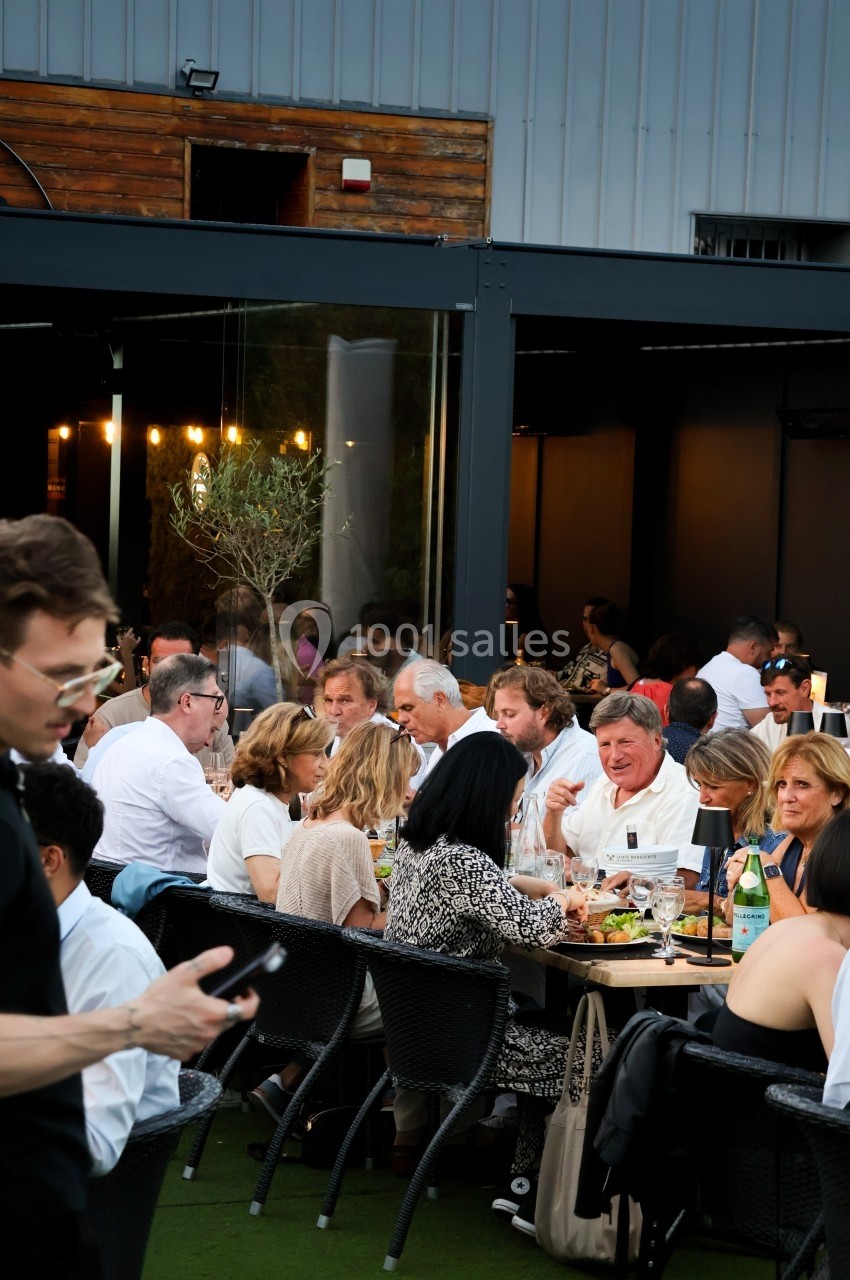 Des personnes dînent en groupe à l'extérieur, autour de grandes tables, dans une ambiance conviviale en soirée.