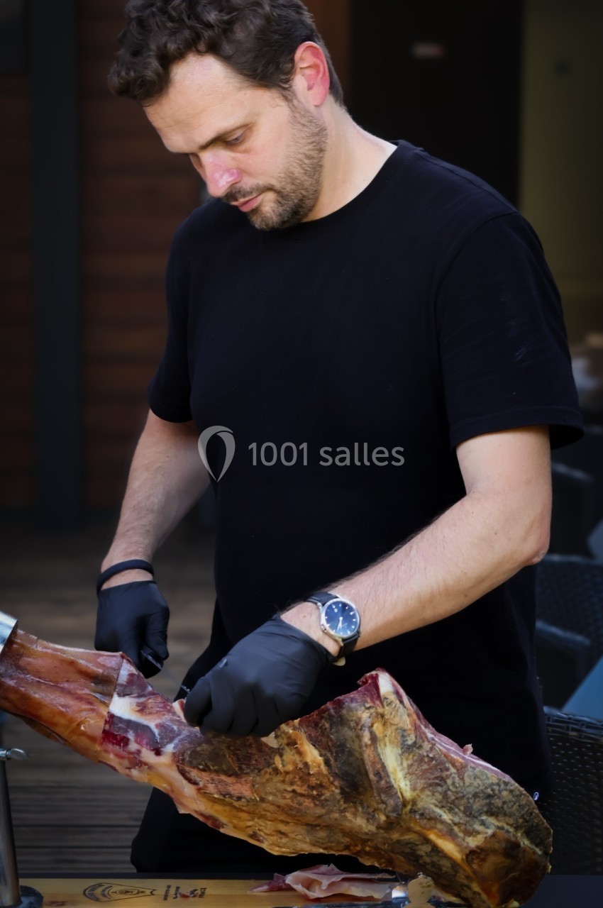 Un homme en t-shirt noir découpe un jambon sur un support, portant des gants noirs, en extérieur.