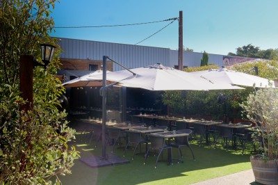 Miniature Location salle Toulouse (Haute-Garonne) - La Cantine d'Horace #18 Terrasse extérieure avec tables et chaises sous des parasols, entourée de verdure et située près d'un bâtiment industriel.