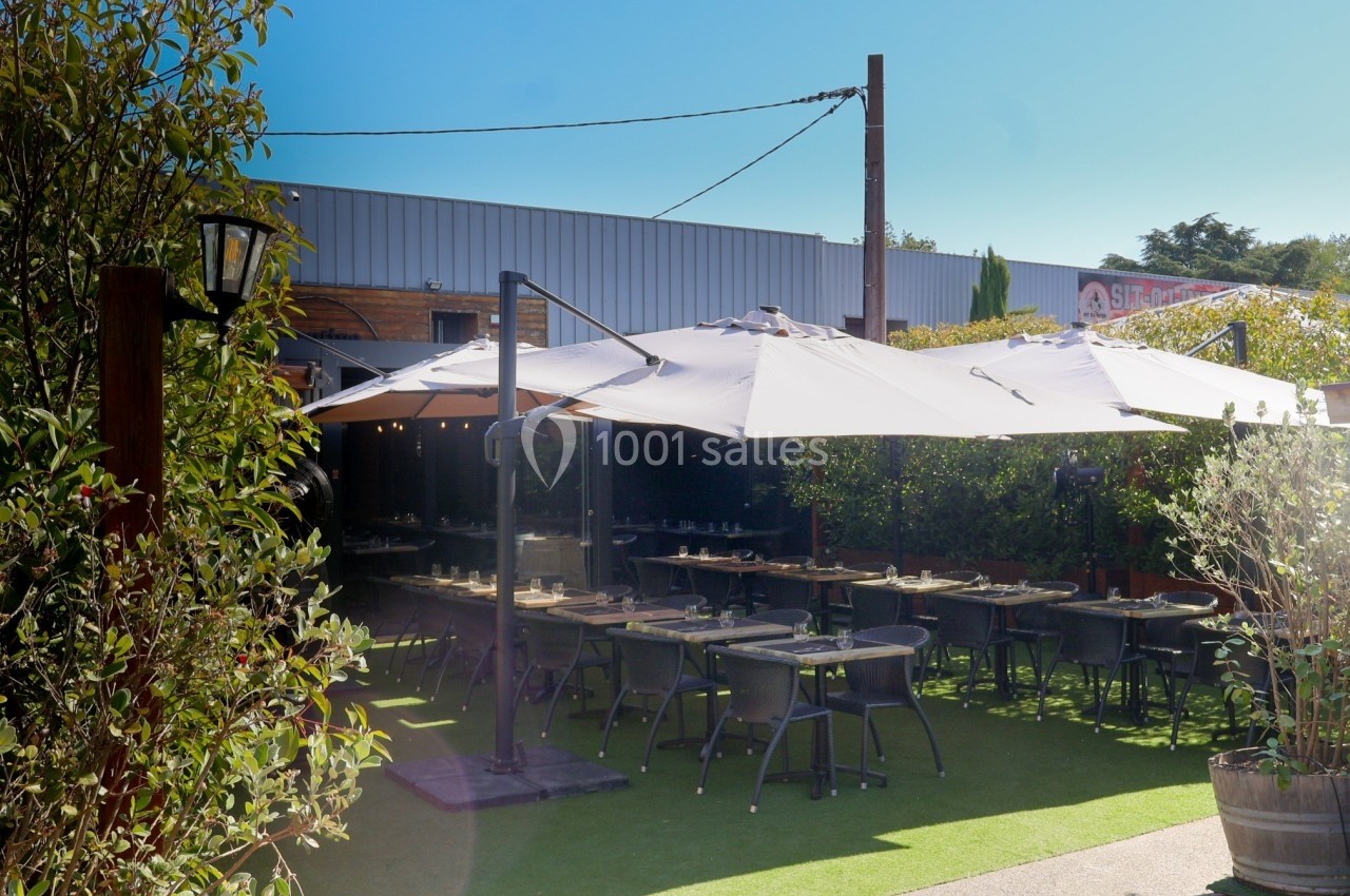 Terrasse extérieure avec tables et chaises sous des parasols, entourée de verdure et située près d'un bâtiment industriel.