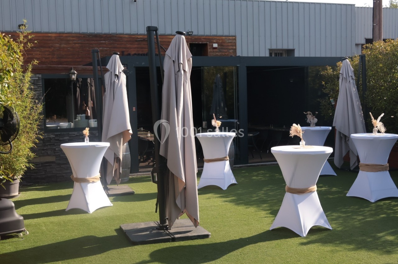 Terrasse aménagée avec des tables hautes drapées de blanc, des parasols fermés et un sol en gazon synthétique.