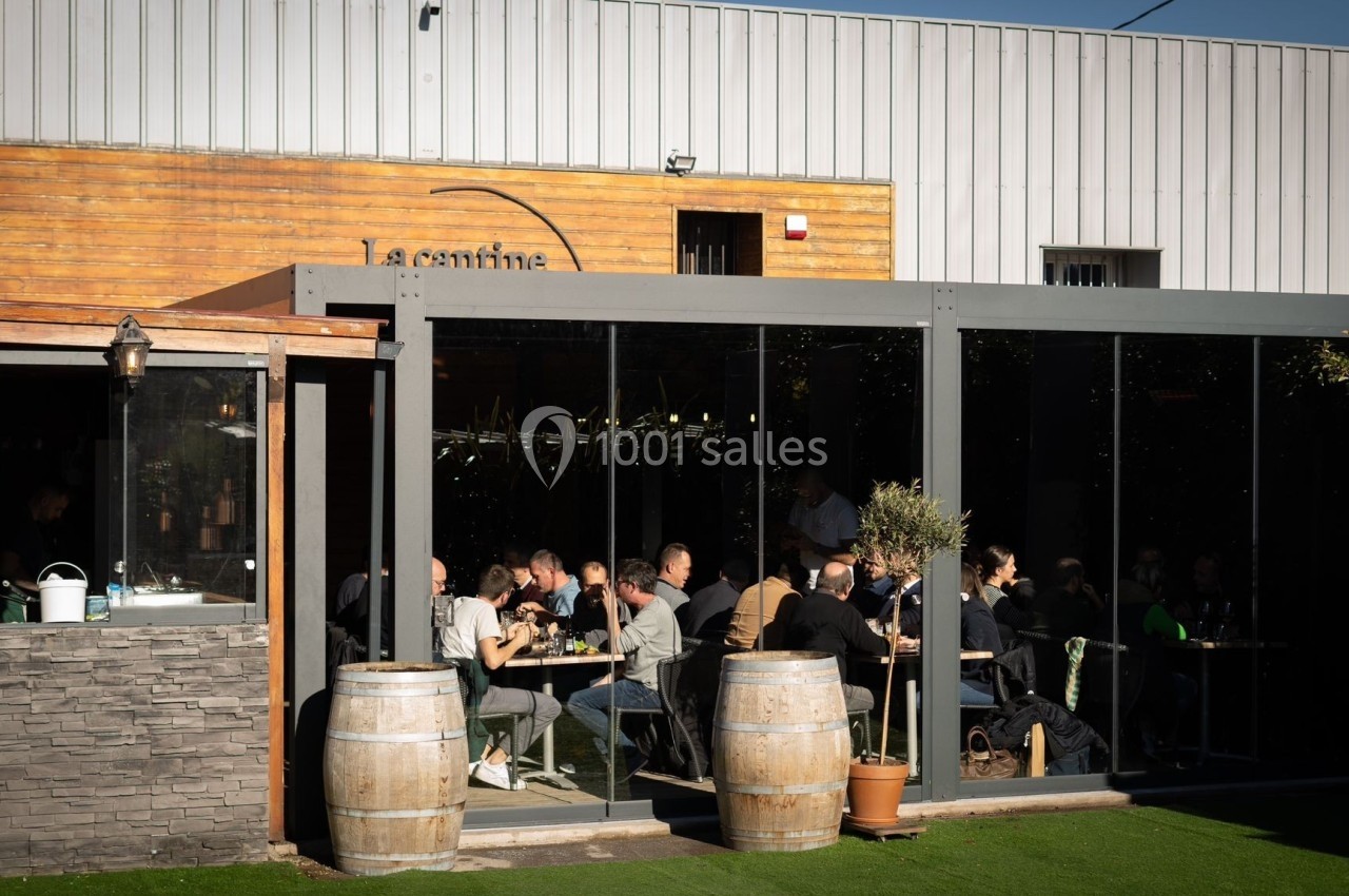 Terrasse extérieure d'un restaurant avec des clients attablés, entourée de barriques et d'une structure vitrée.