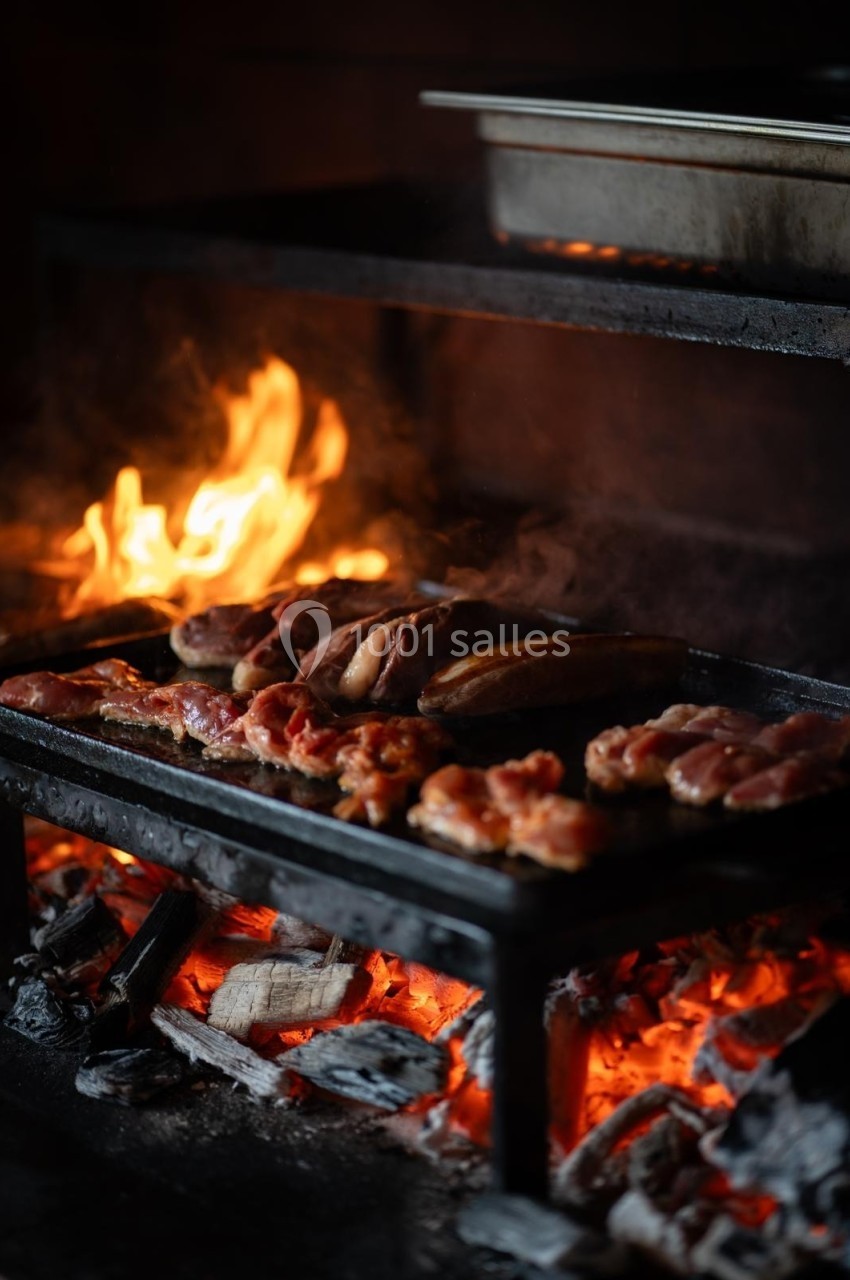 Viande grillant sur une grille au-dessus de braises ardentes, avec des flammes en arrière-plan.