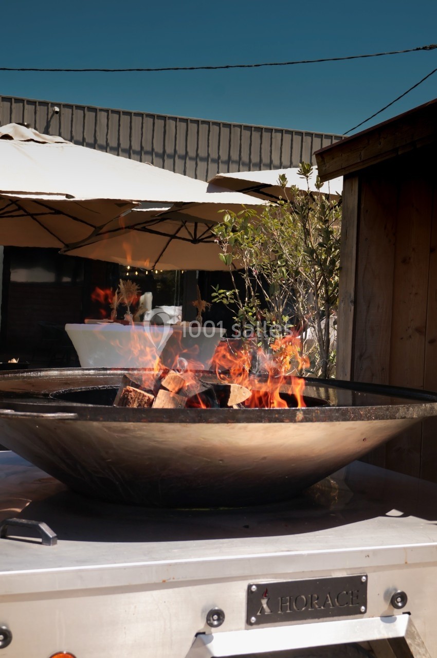 Brasero allumé avec des flammes vives, placé sur une terrasse extérieure près d'arbustes et de parasols.