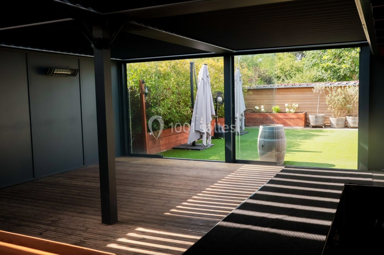 Vue d'une terrasse couverte avec sol en bois, ouverte sur un jardin avec pelouse, parasol et plantes en pots.