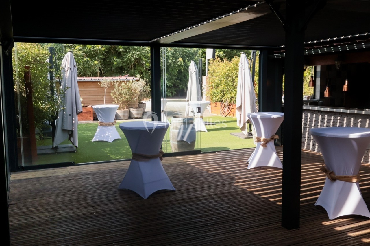 Terrasse aménagée avec tables hautes habillées de nappes blanches, parasols et verdure en arrière-plan.