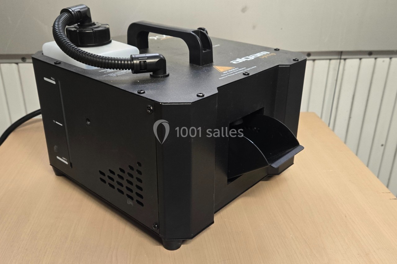 Machine à fumée noire posée sur une table en bois, avec réservoir visible et sortie de fumée orientée vers l'avant.
