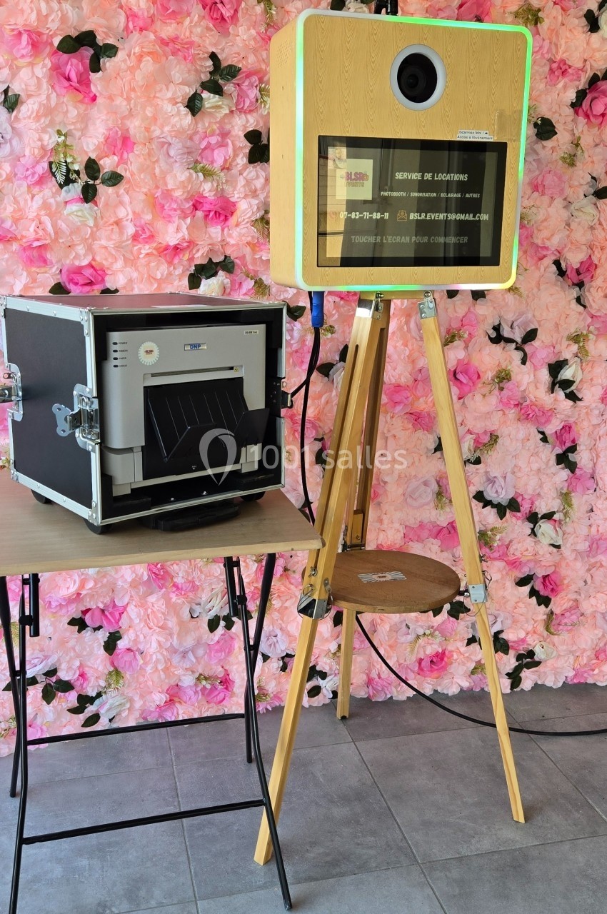 Borne photo en bois sur trépied avec imprimante à côté, devant un mur floral rose et un éclairage studio.