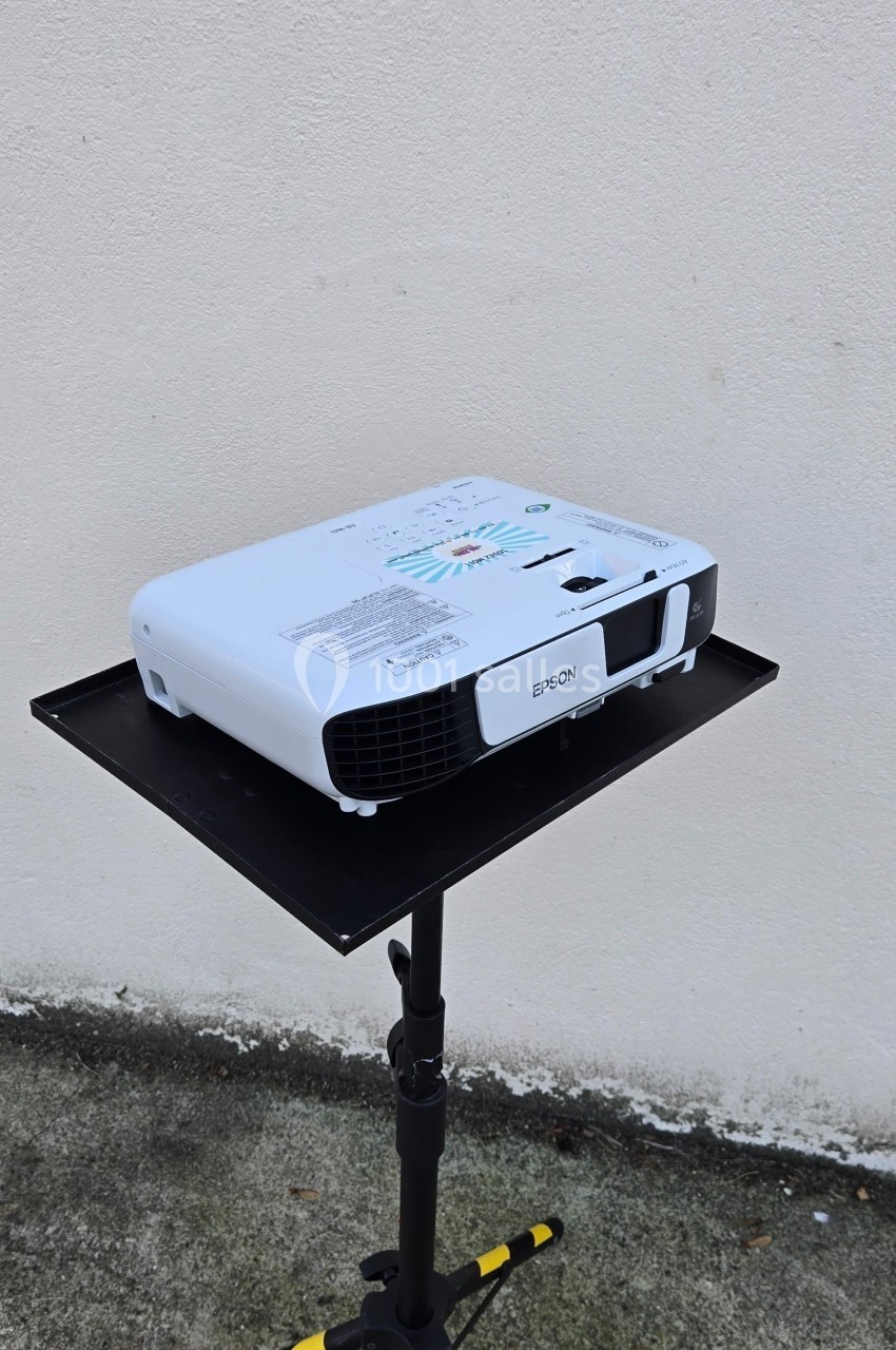 Projecteur blanc posé sur un support noir devant un mur clair, avec un sol en béton visible.