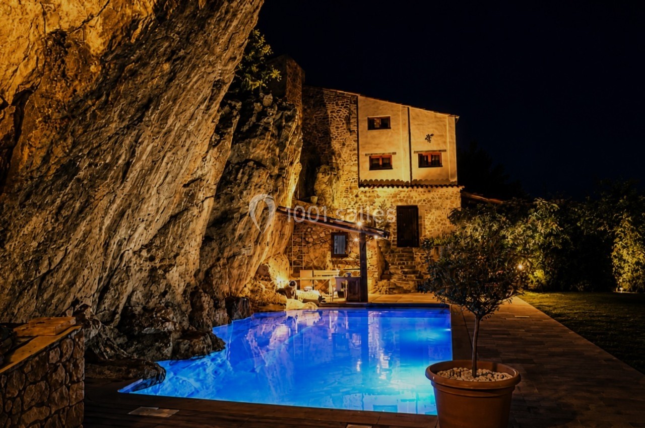 Piscine éclairée de nuit au pied d'une falaise, avec une maison en pierre illuminée en arrière-plan.