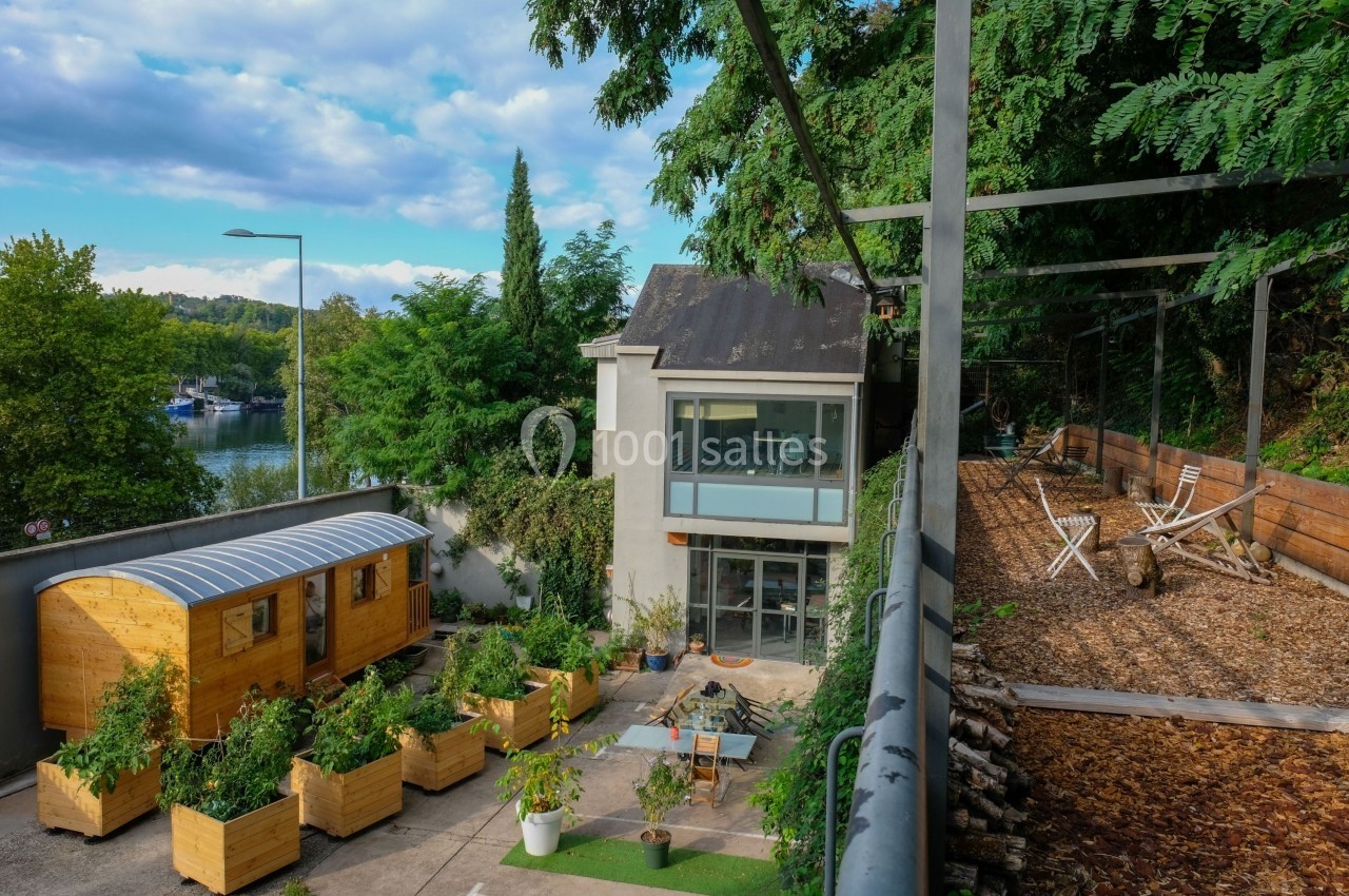 Vue d'une maison moderne avec terrasse, jardin aménagé, potager en bacs et roulotte en bois, entourée de verdure.