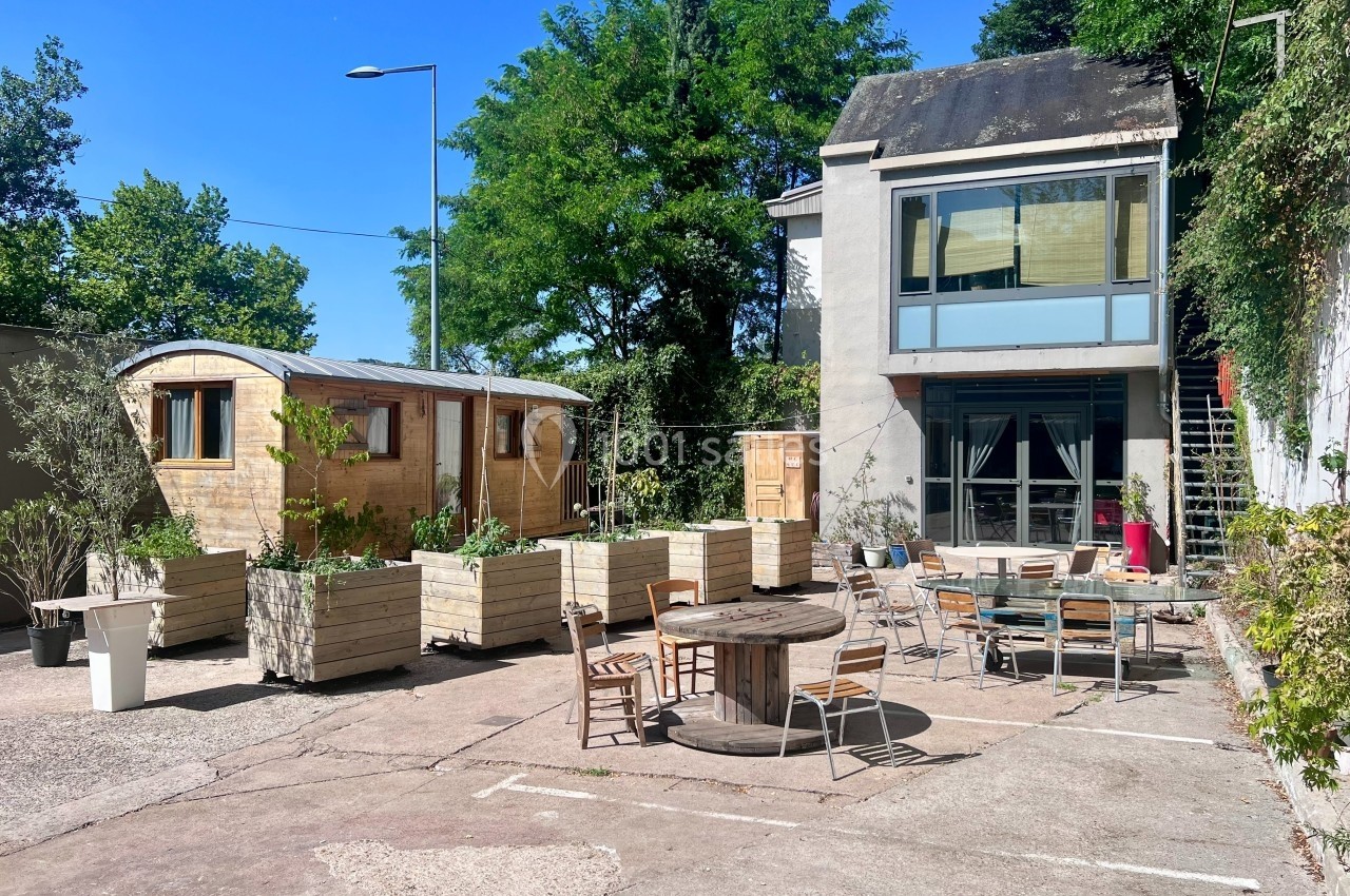 Cour extérieure avec tables, chaises, jardinières en bois, roulottes et bâtiment moderne entouré de verdure.