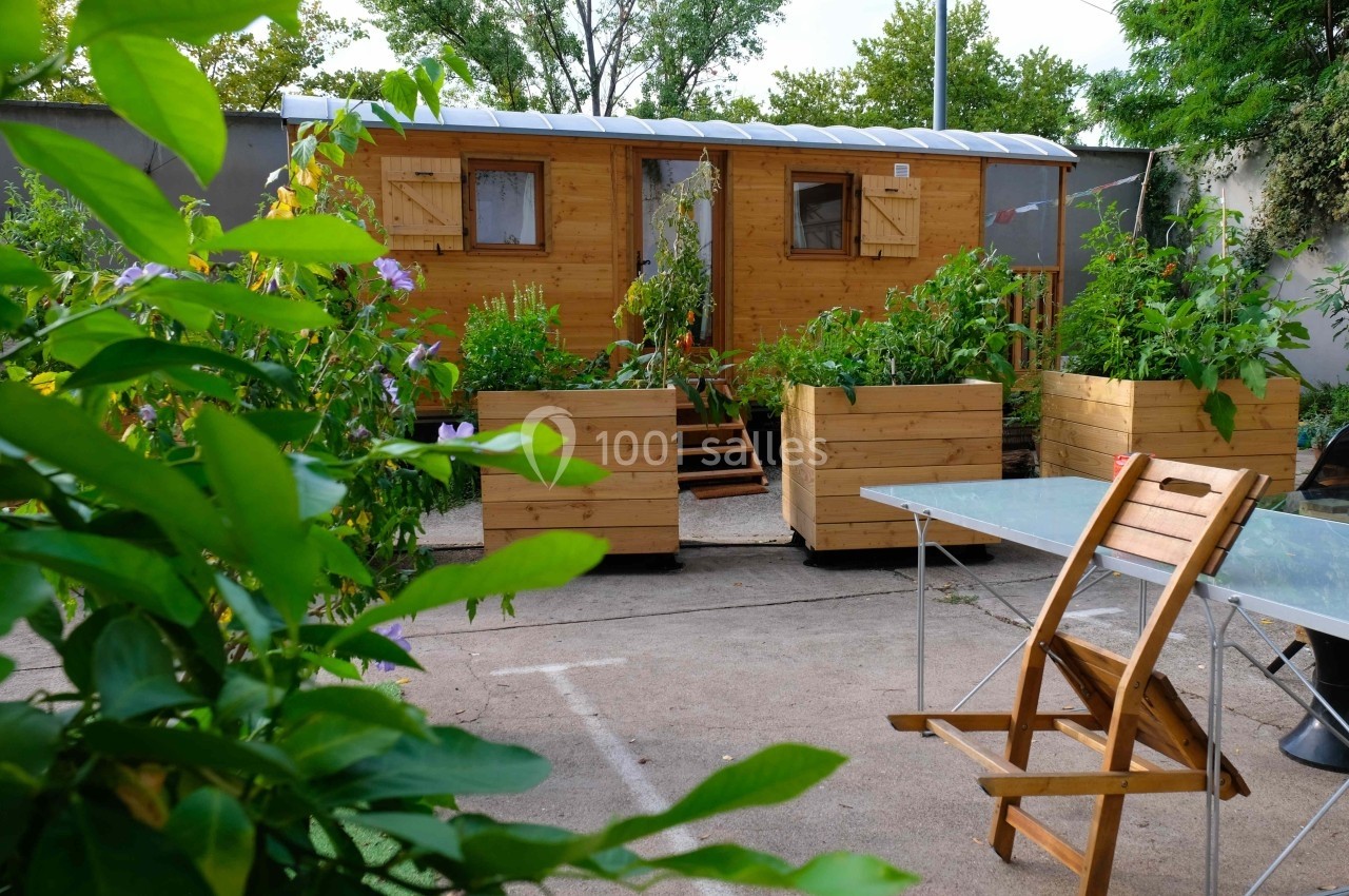 Cabane en bois entourée de jardinières avec des plantes, située dans une cour avec une table et une chaise au premier plan.