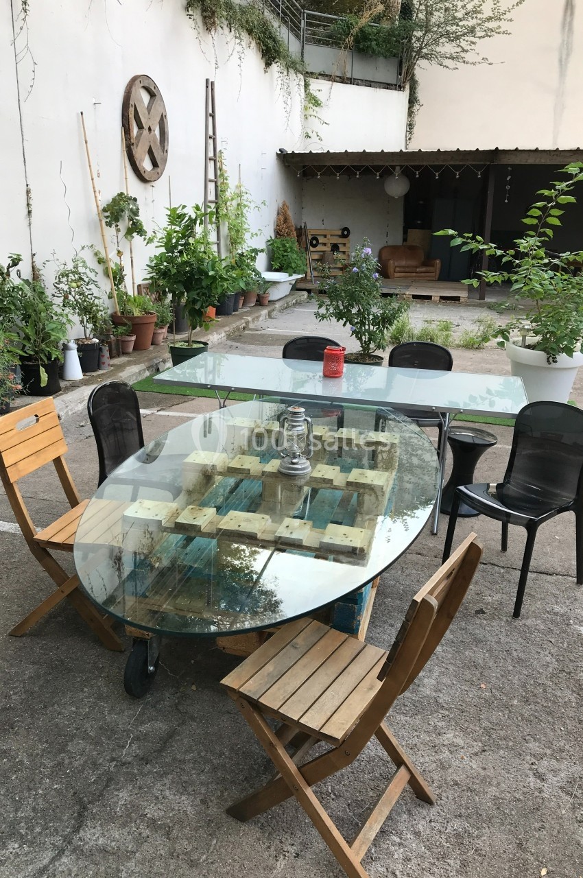Table en verre entourée de chaises en bois et en métal, située dans une cour avec des plantes en pots et un abri au fond.