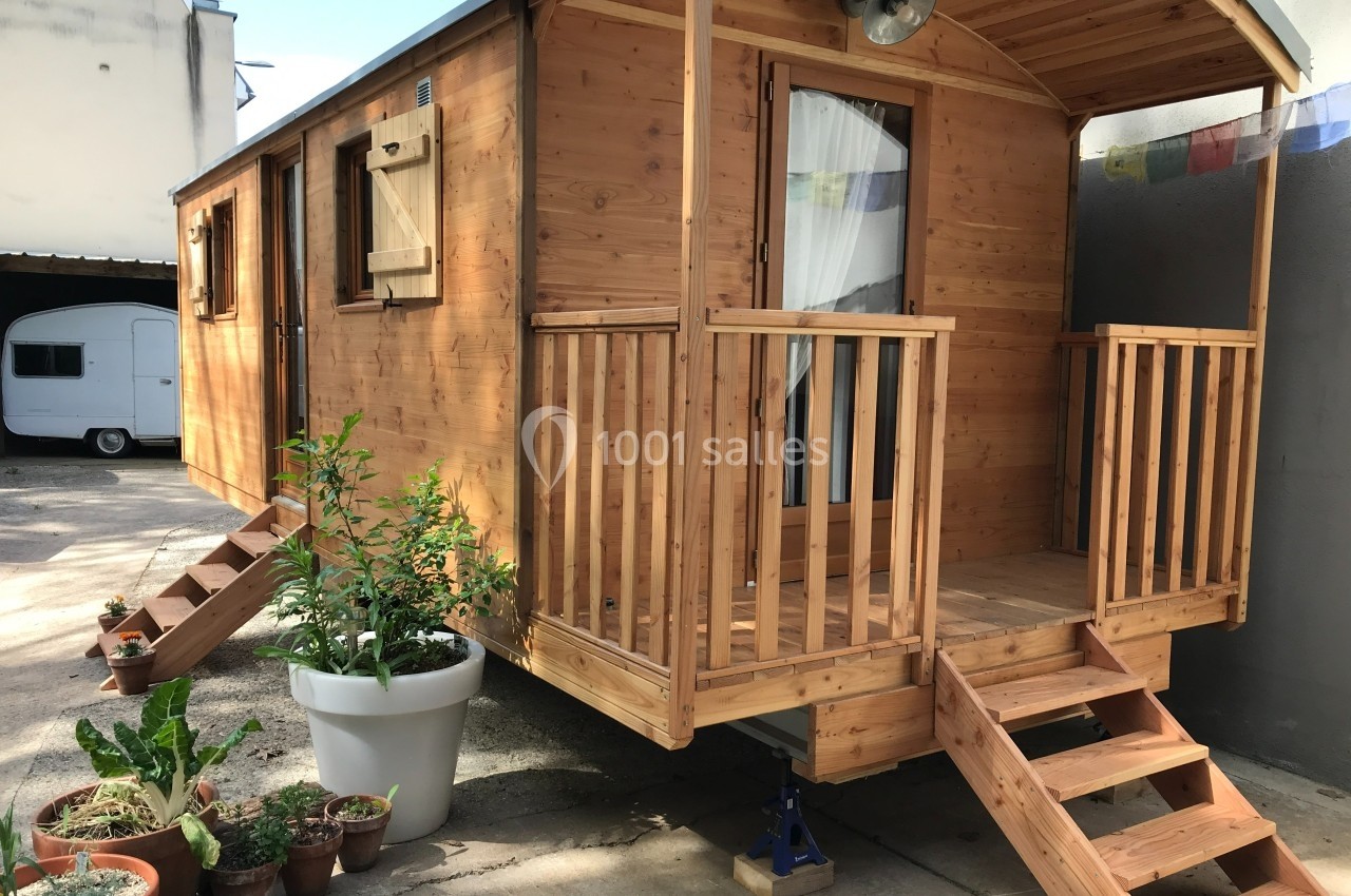 Roulotte en bois avec terrasse et escaliers, entourée de plantes en pots, stationnée dans une cour.