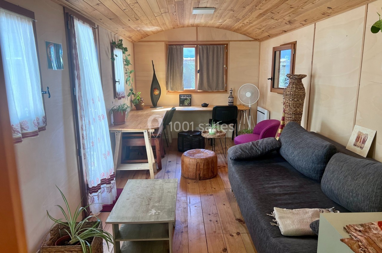 Intérieur d'une petite pièce en bois avec canapé, bureau, plantes et lumière naturelle entrant par les fenêtres.