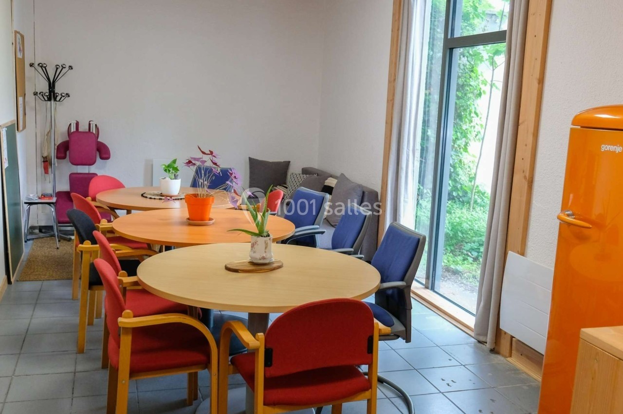 Salle de détente avec tables rondes, chaises colorées, canapé, plantes et une fenêtre donnant sur un espace vert.