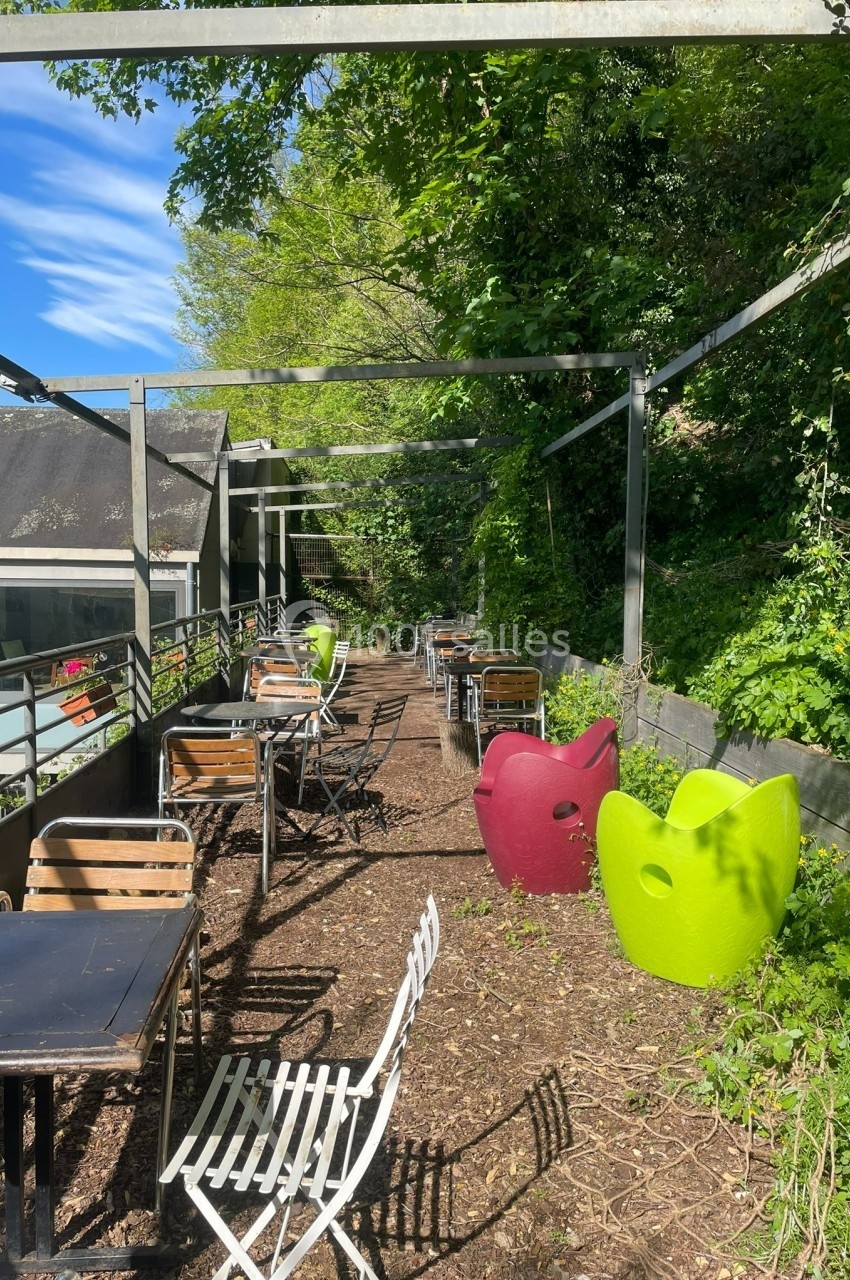 Terrasse extérieure avec tables, chaises et fauteuils colorés, entourée de végétation et partiellement ombragée.