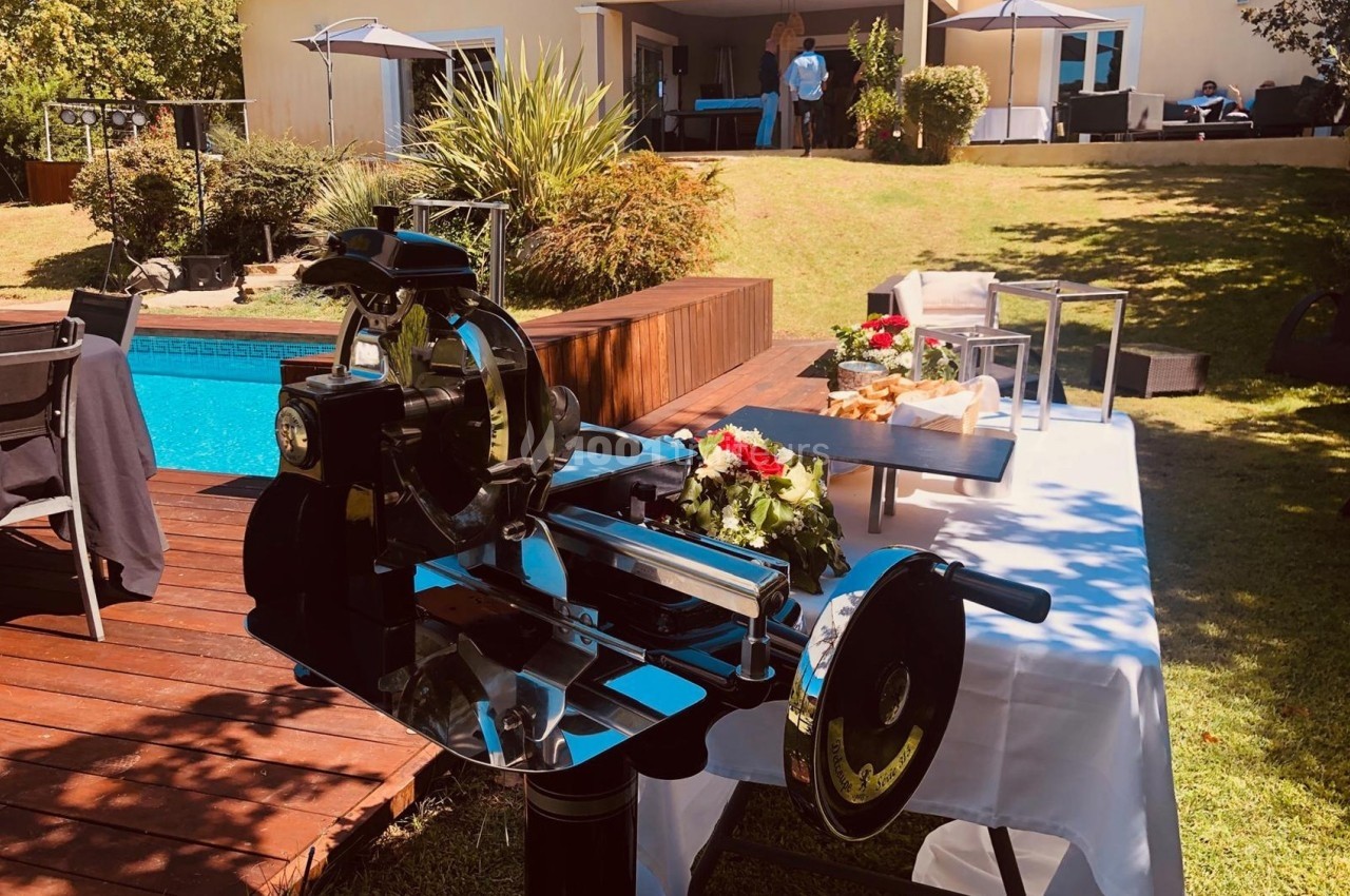 Trancheuse à jambon installée près d'une table dressée dans un jardin avec piscine et maison en arrière-plan.