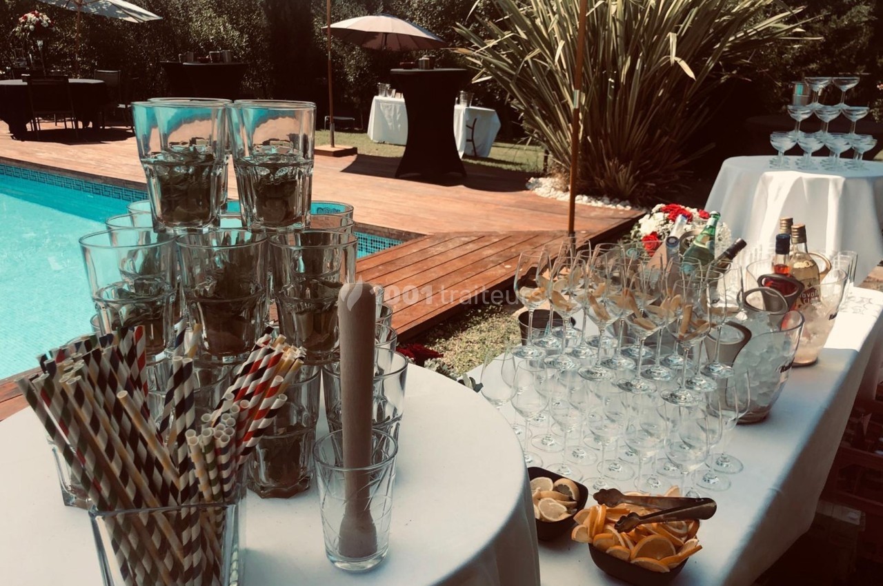 Table dressée en extérieur près d'une piscine, avec verres, pailles, citrons et bouteilles pour un service de boissons.