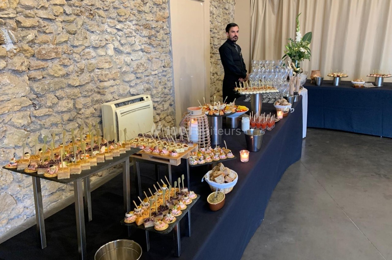 Buffet de réception avec verrines, amuse-bouches et boissons, disposé sur des tables noires dans une salle en pierre.