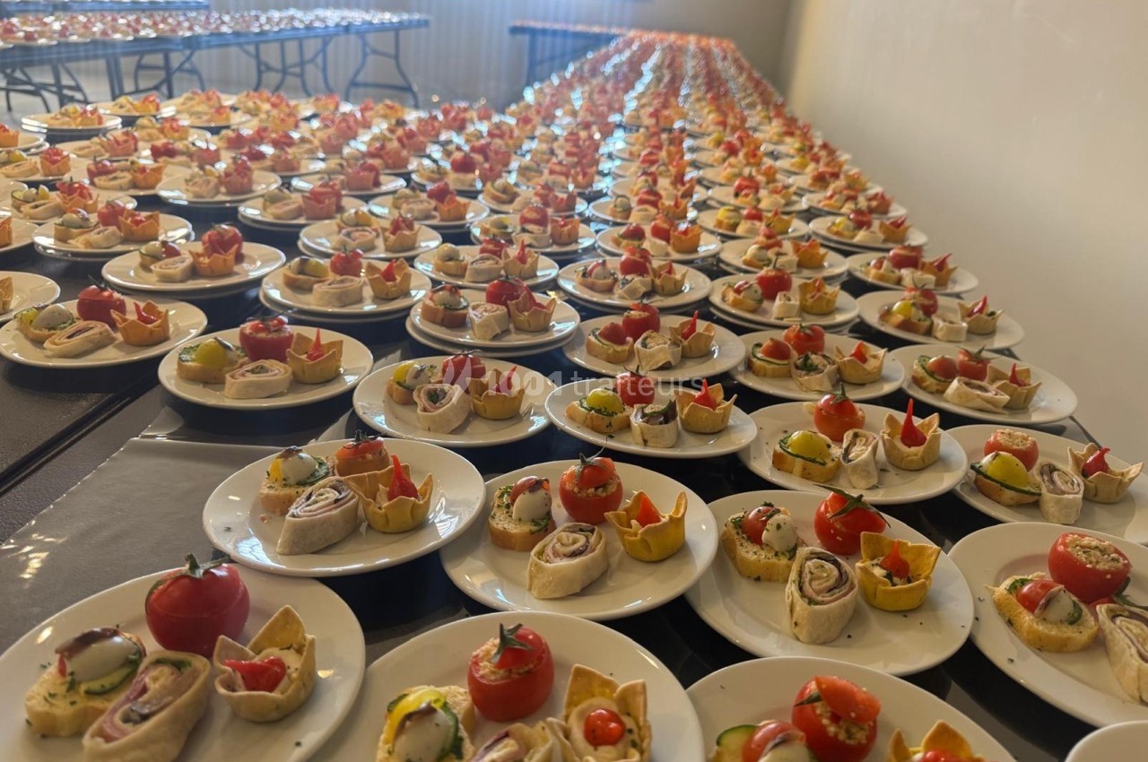 Assiettes alignées contenant des amuse-bouches variés, avec tomates cerises, wraps et tartelettes, dans une grande salle.