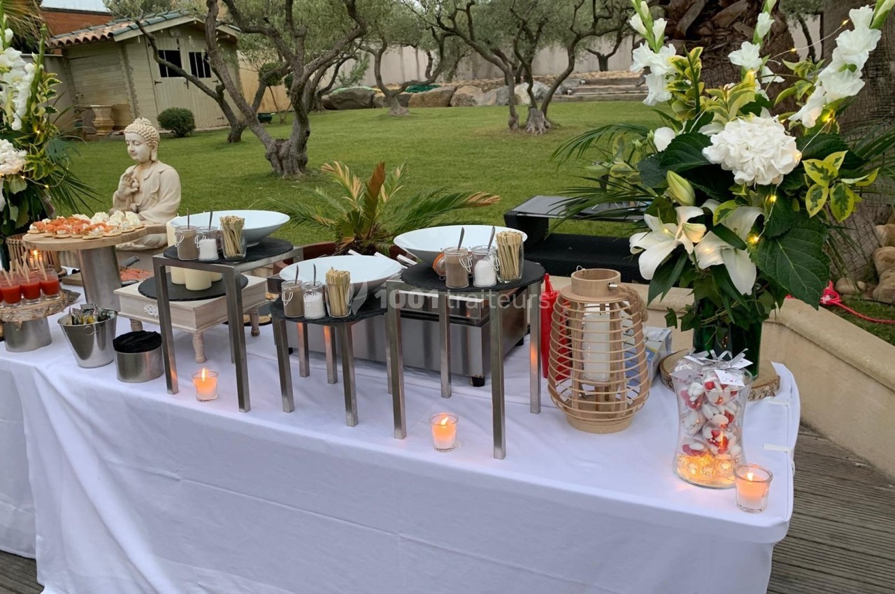 Buffet extérieur élégant avec plats variés, décoré de fleurs blanches, bougies et statue de Bouddha dans un jardin.