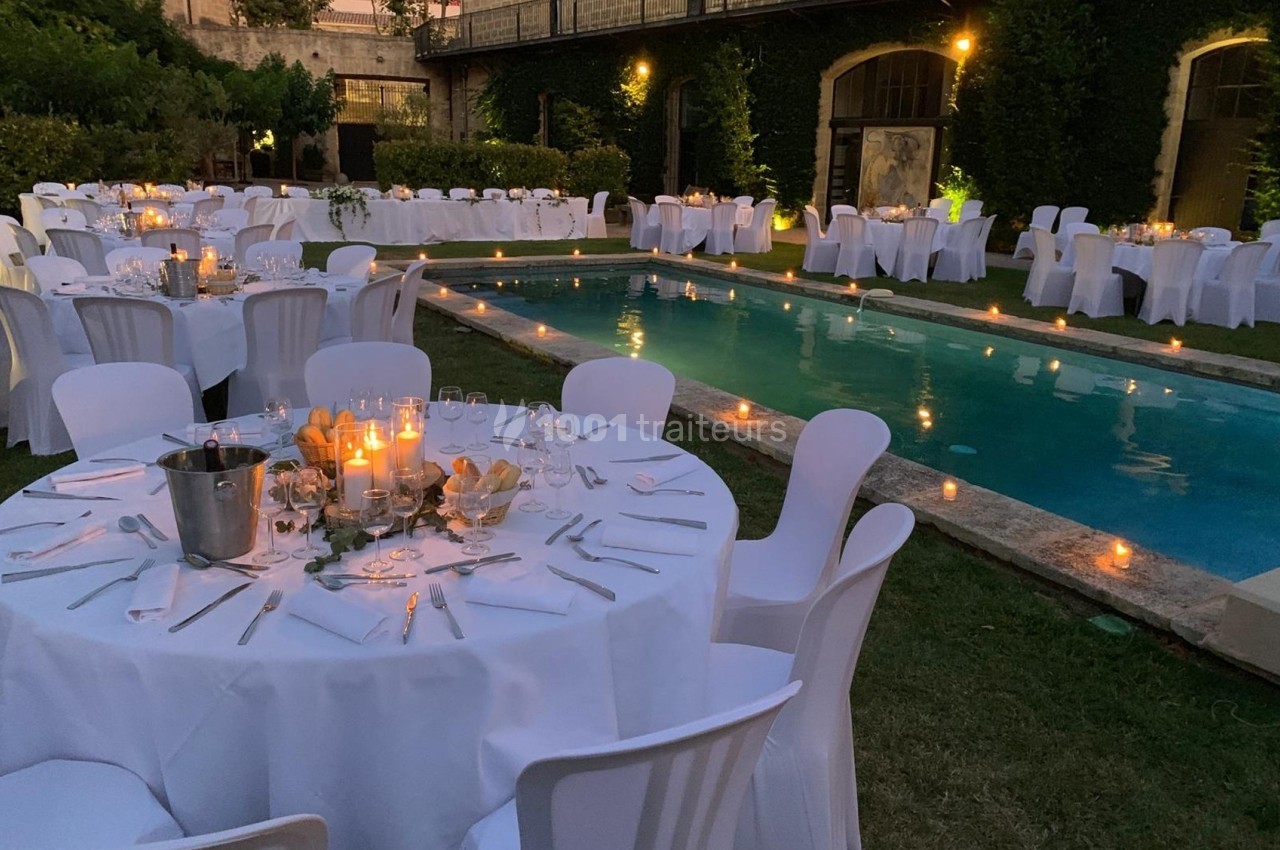 Tables dressées avec nappes blanches autour d'une piscine éclairée par des bougies dans un jardin en soirée.