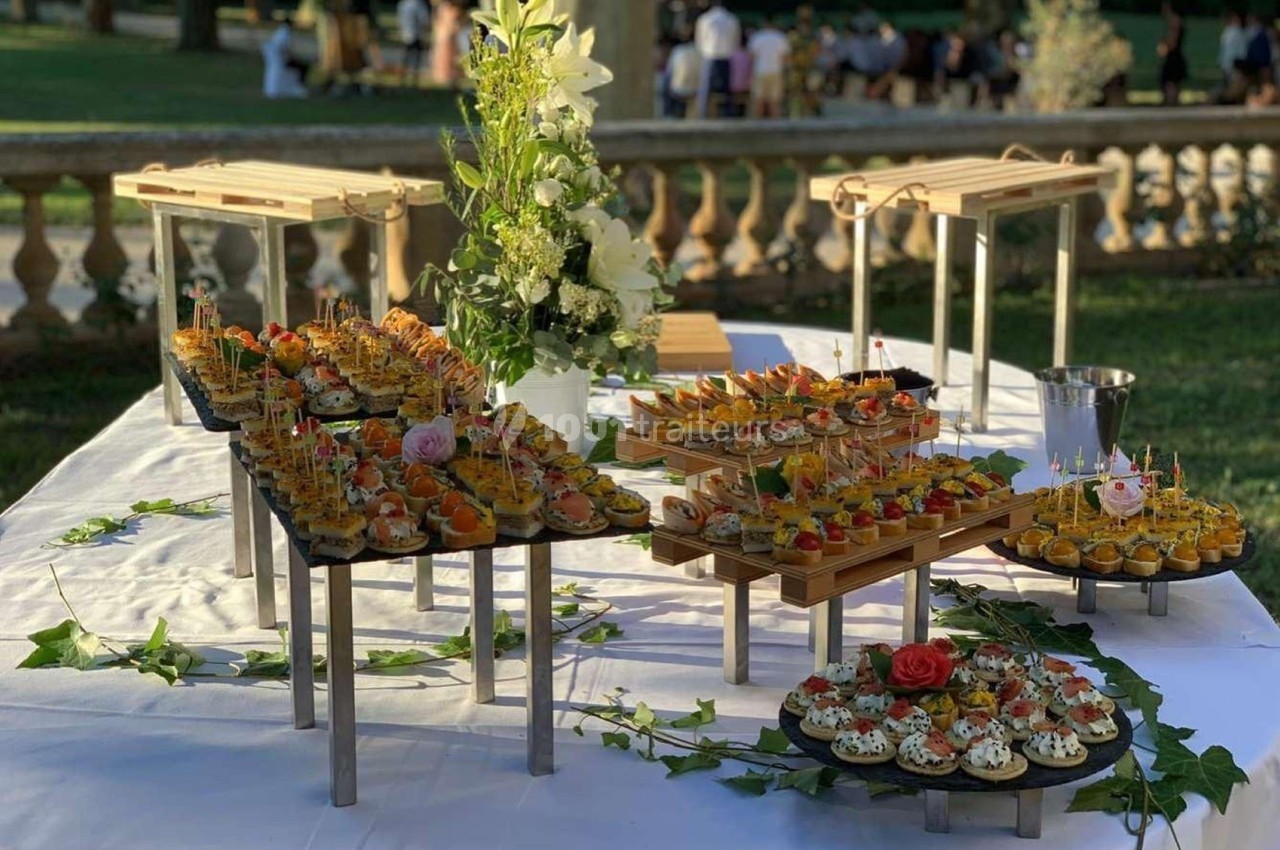Table dressée en extérieur avec des plateaux de canapés variés, décorée de fleurs et de feuillage.