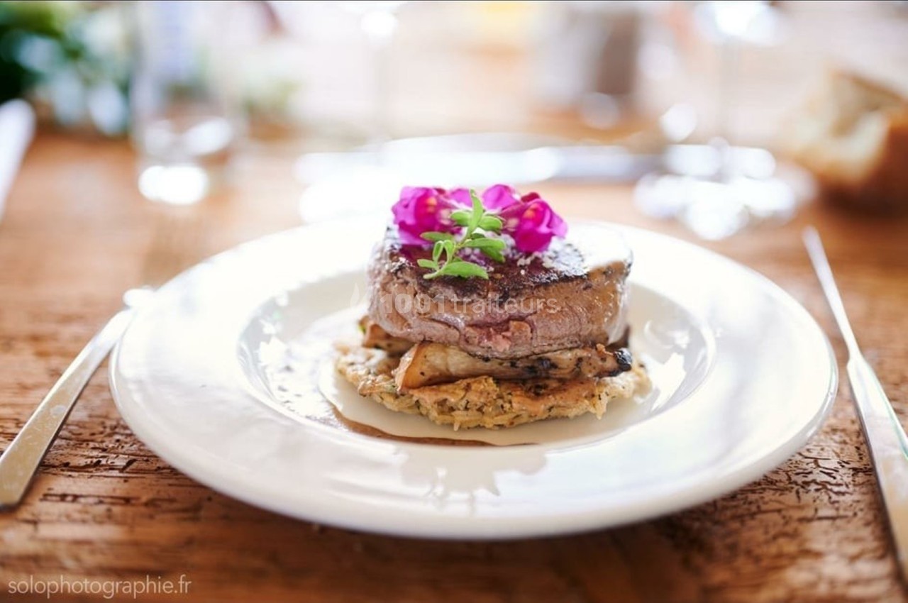 Assiette avec un filet de viande sur un lit de légumes et d'herbes, décoré de fleurs comestibles, sur une table en bois.