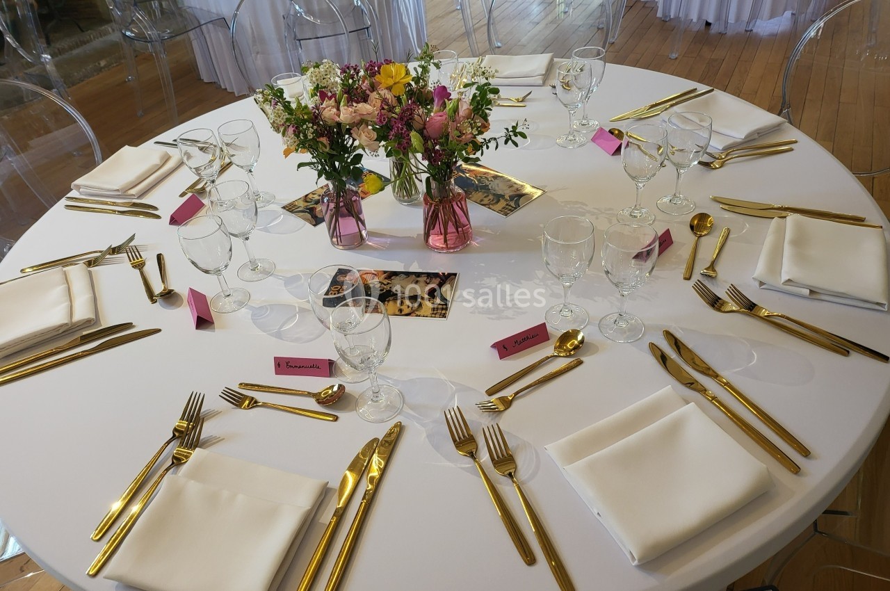 Table ronde dressée avec nappes blanches, couverts dorés, verres, serviettes pliées et centre de table floral coloré.