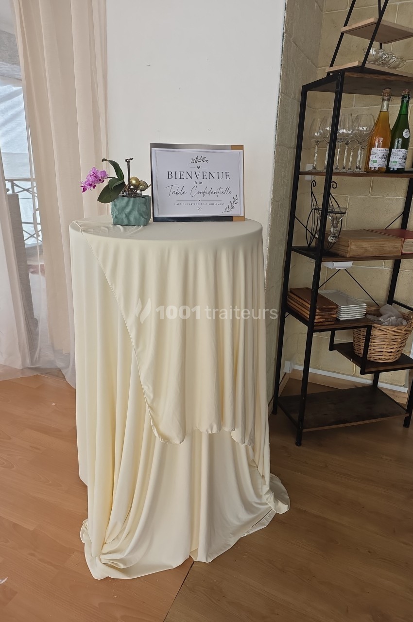 Table haute recouverte d'une nappe beige, décorée d'une orchidée et d'un panneau de bienvenue, près d'une étagère.