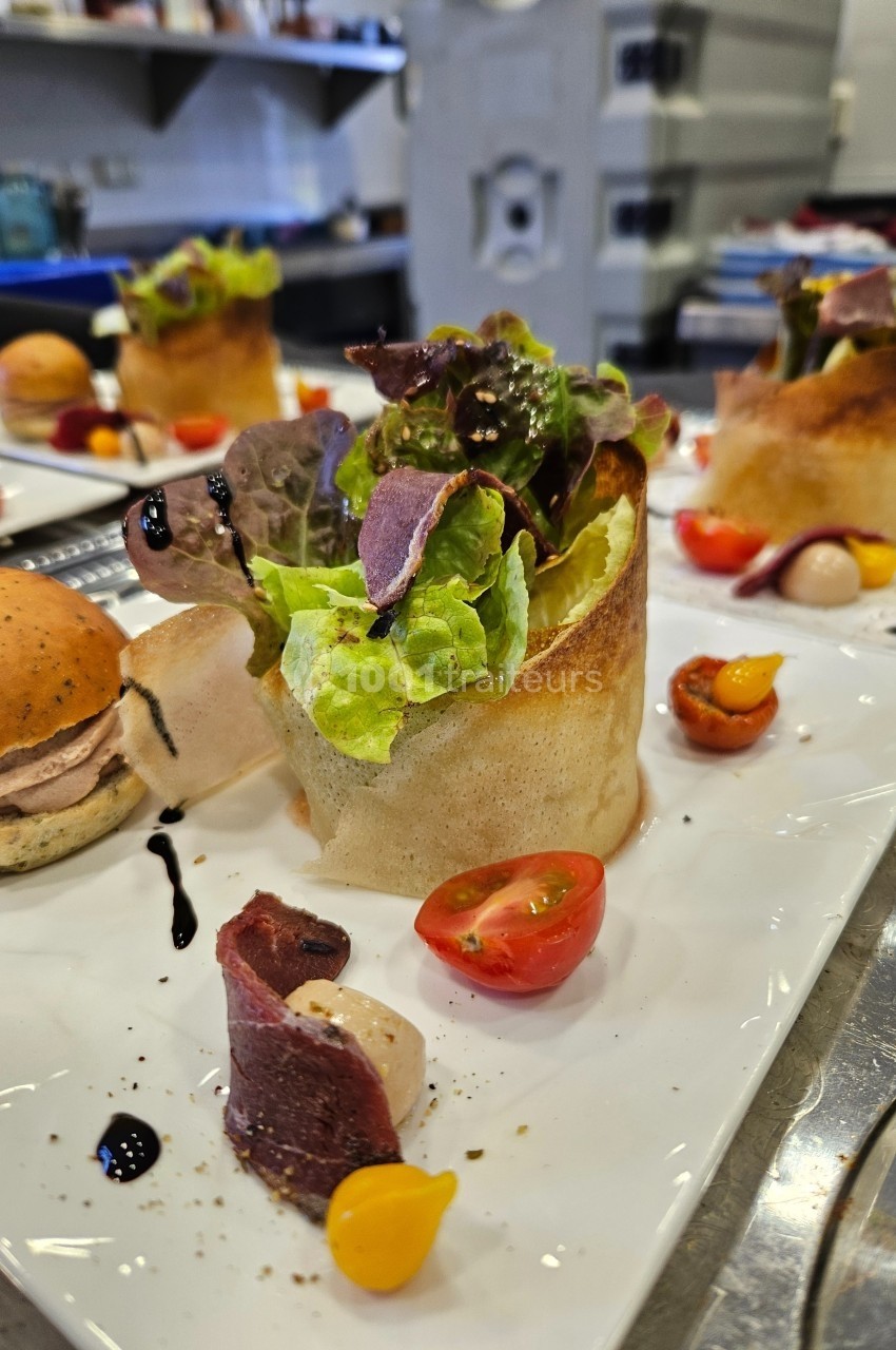 Assiette gastronomique avec salade, tomates cerises, viande séchée et sauce balsamique, présentée sur une table de cuisine.