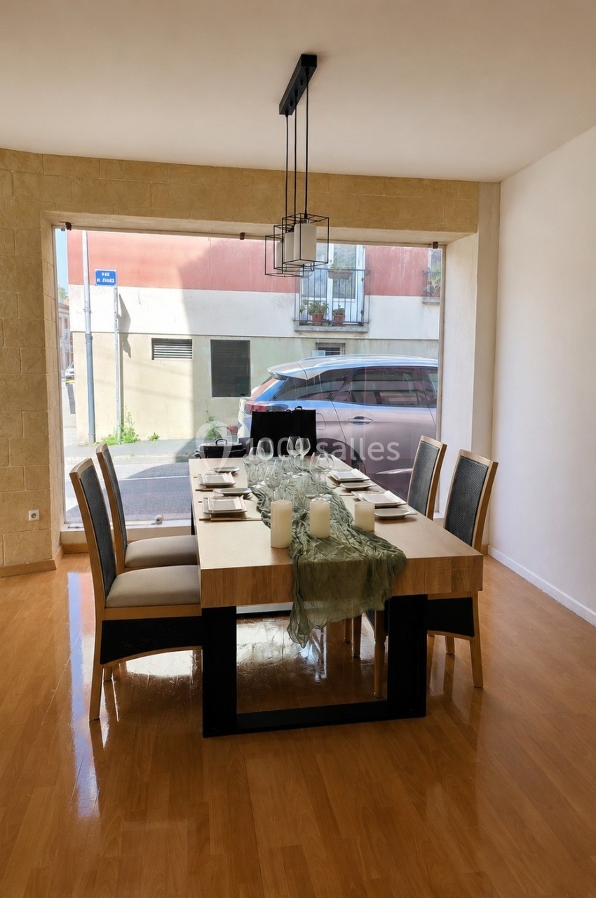 Salle à manger lumineuse avec une grande table en bois, six chaises, des bougies et une nappe décorative, vue sur la rue.