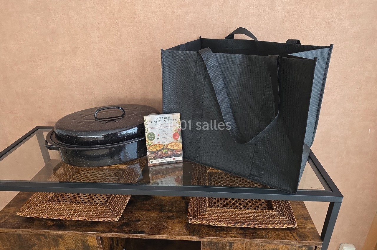 Cocotte noire avec couvercle, sac réutilisable noir et dépliant posés sur une table en verre avec sets en osier.