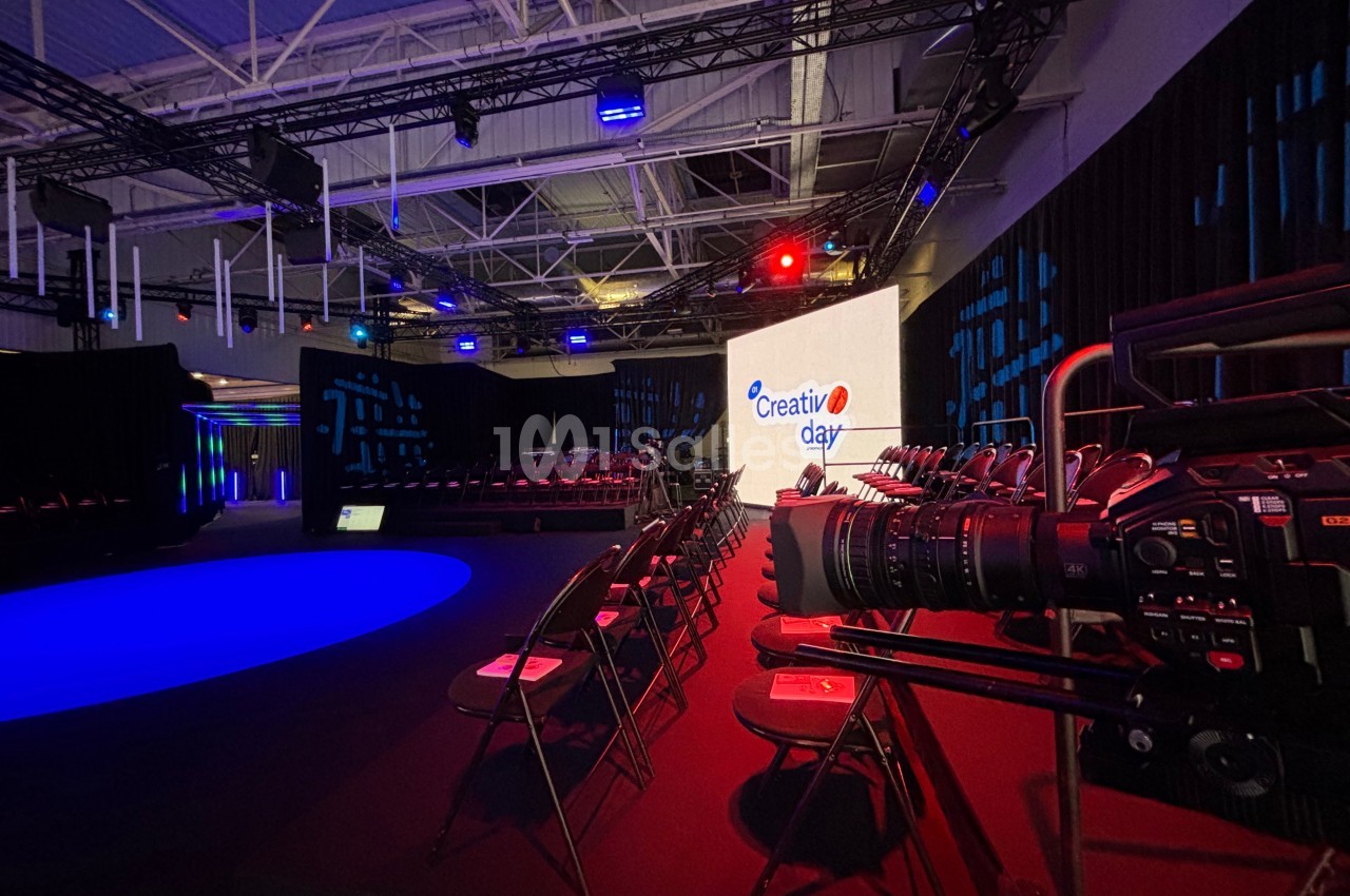 Salle de conférence avec éclairage coloré, écran affichant ’Creativ Day’, rangées de chaises et caméra sur trépied.
