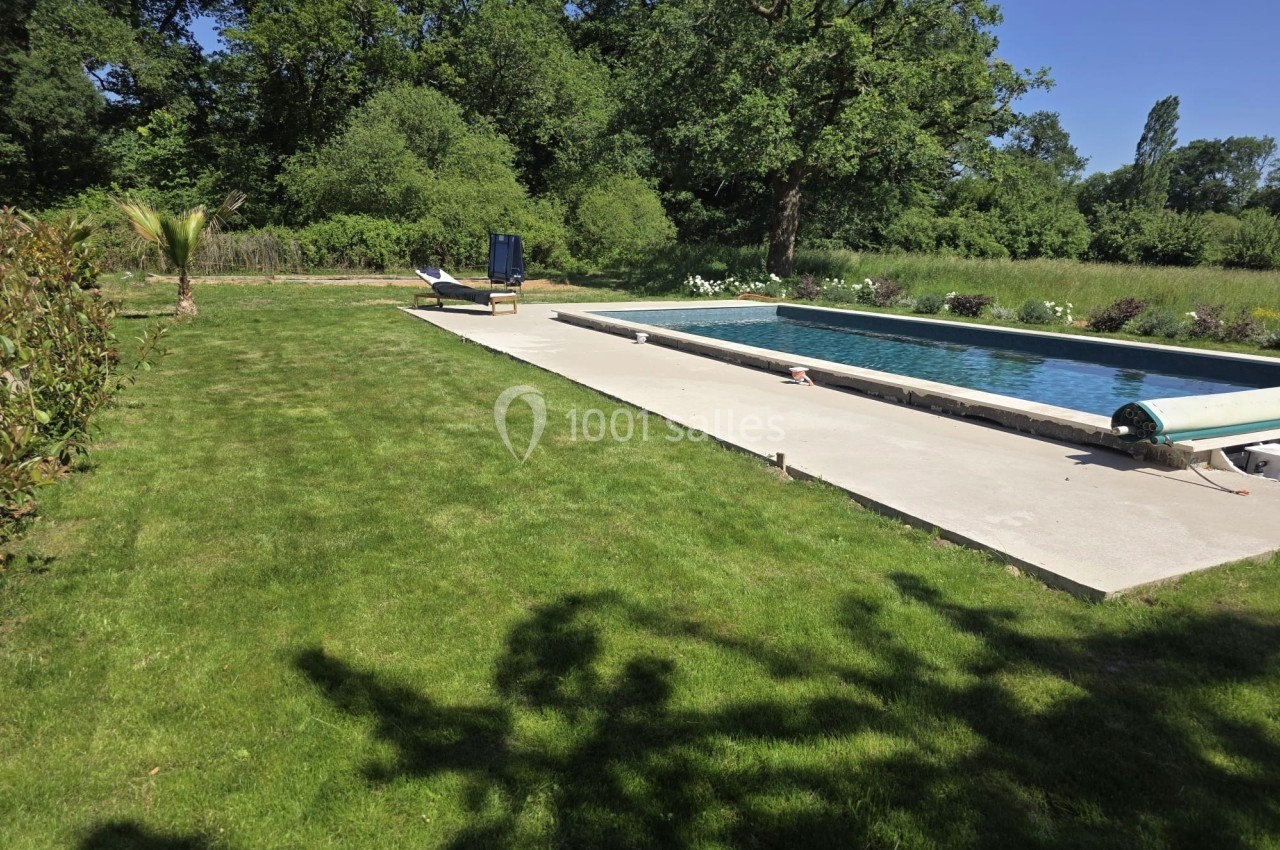 Piscine extérieure rectangulaire entourée de pelouse et d'arbres, avec un transat et un parasol à l'arrière-plan.