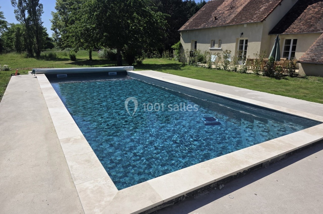 Piscine rectangulaire avec eau claire entourée d'une terrasse en béton, située près d'une maison et d'un jardin verdoyant.