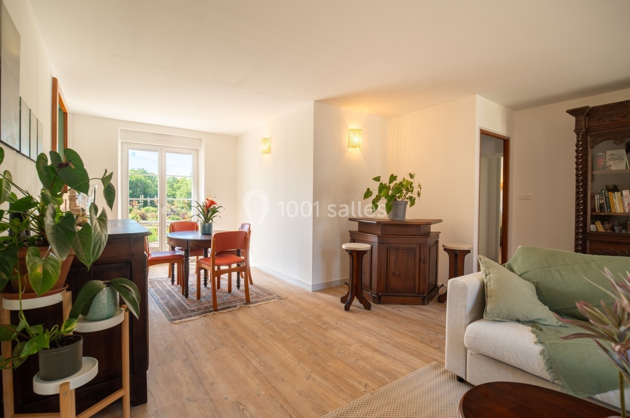 Salon lumineux avec parquet, meubles en bois, plantes vertes et vue sur un extérieur verdoyant par une fenêtre.