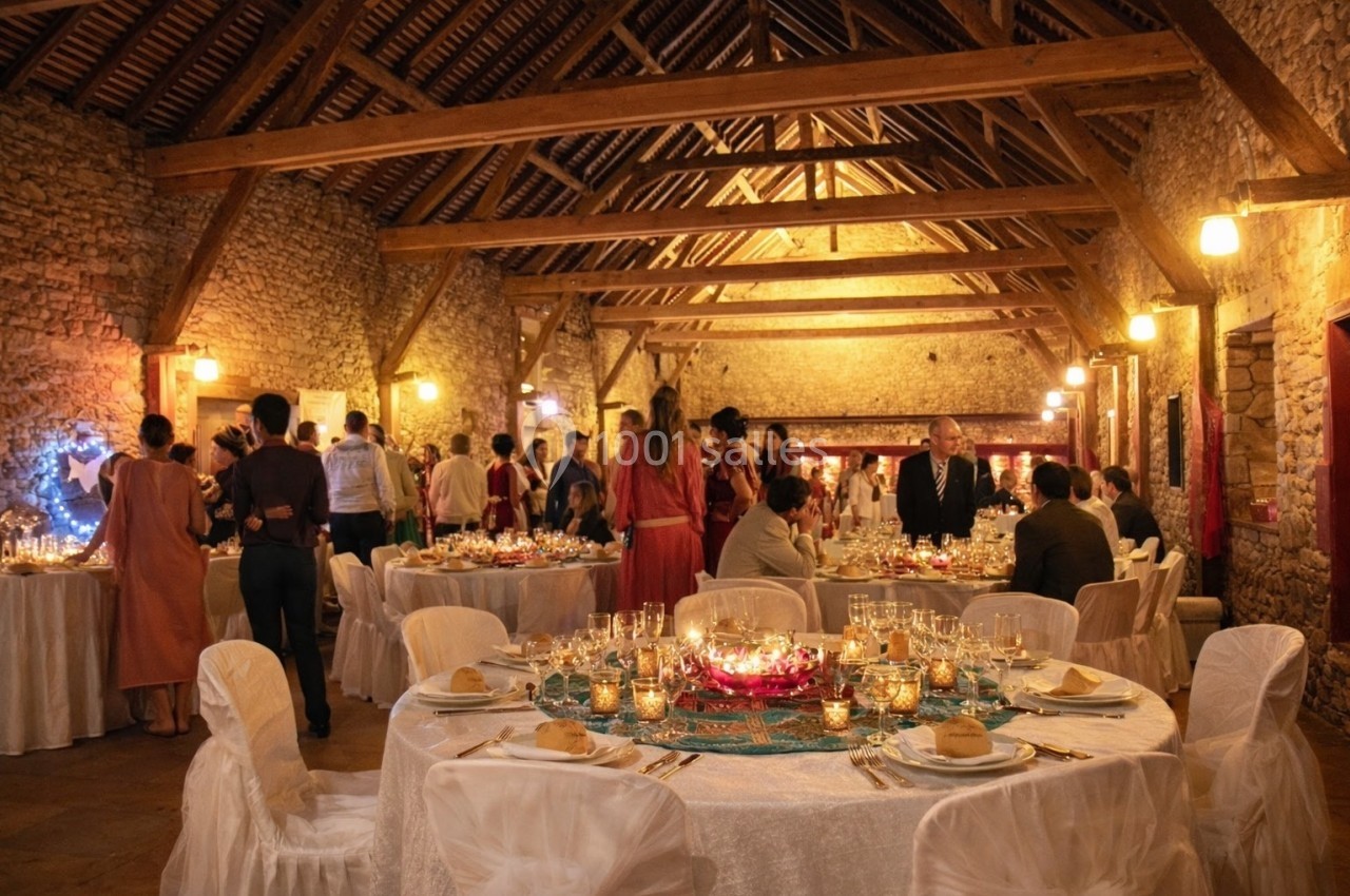 Salle de réception en pierre avec des invités assis et debout, tables décorées de bougies et vaisselle élégante.