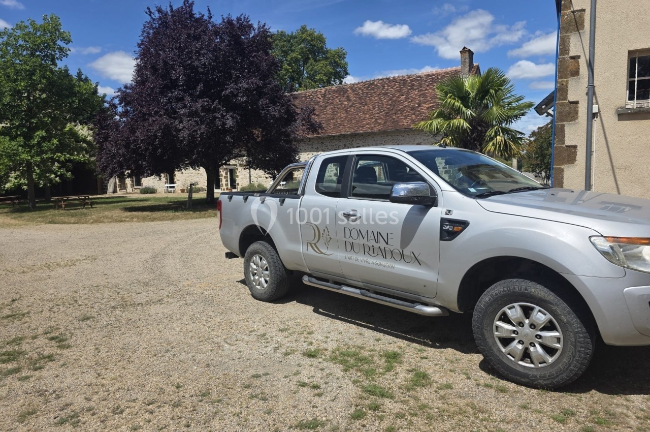Pick-up gris stationné devant un bâtiment en pierre avec un toit en tuiles, entouré d'arbres et d'herbe.