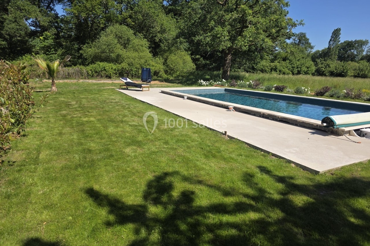 Piscine extérieure entourée de pelouse et de végétation, avec transat et parasol sous un ciel dégagé.