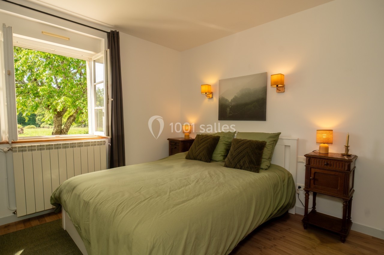 Chambre lumineuse avec lit double, draps verts, tables de chevet en bois, lampes allumées et fenêtre ouverte sur un arbre.