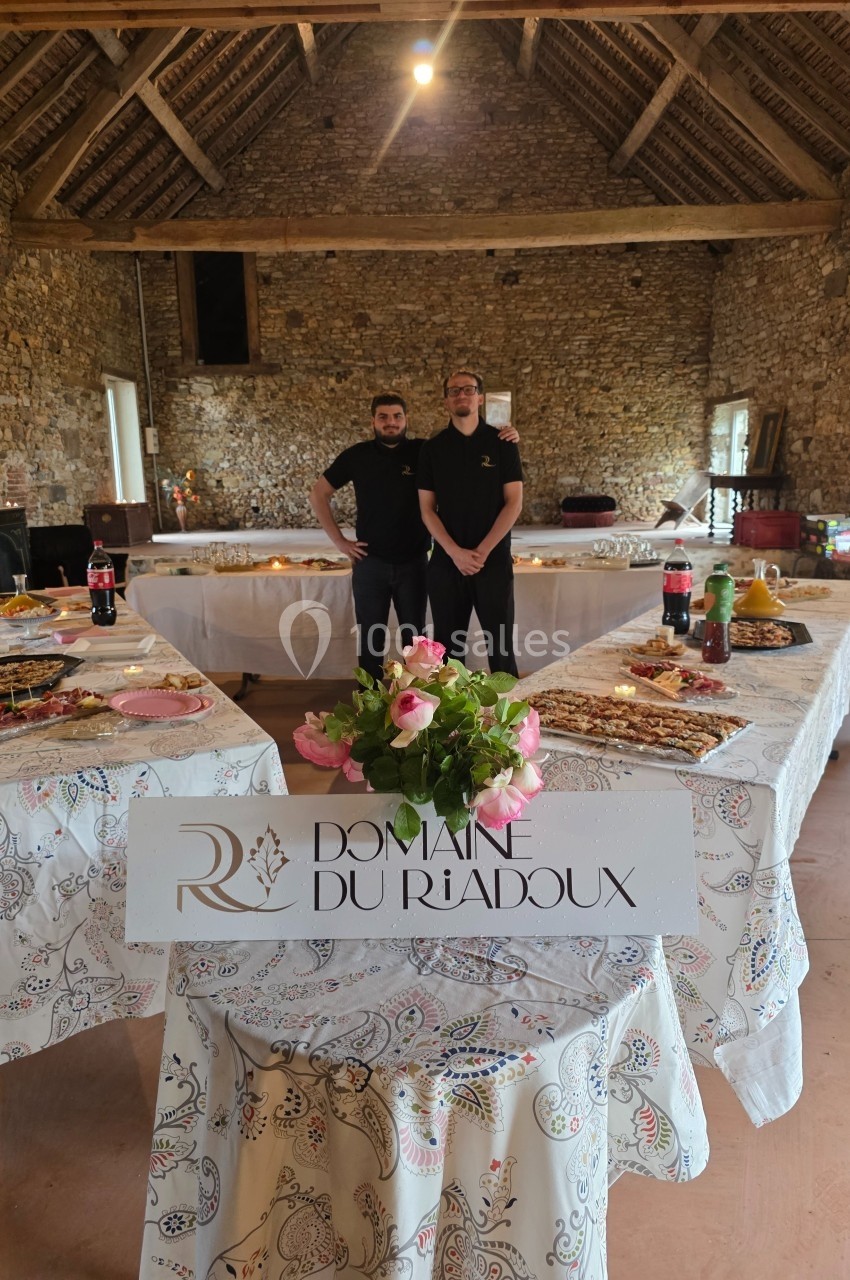 Deux personnes posent dans une salle en pierre avec des tables dressées pour un buffet, décorées de nappes et de fleurs.