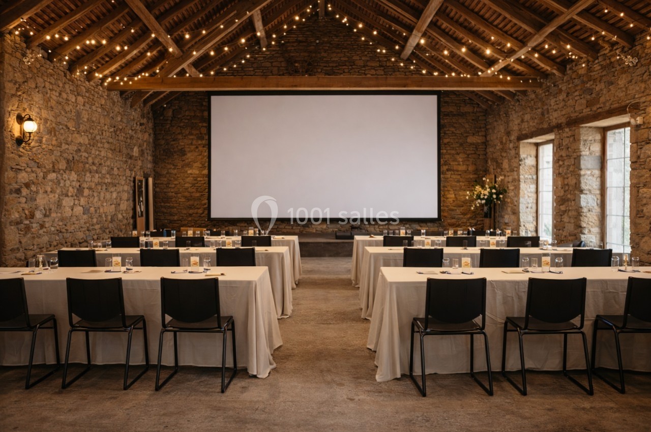 Salle de conférence en pierre avec tables, chaises noires, écran de projection et éclairage suspendu chaleureux.