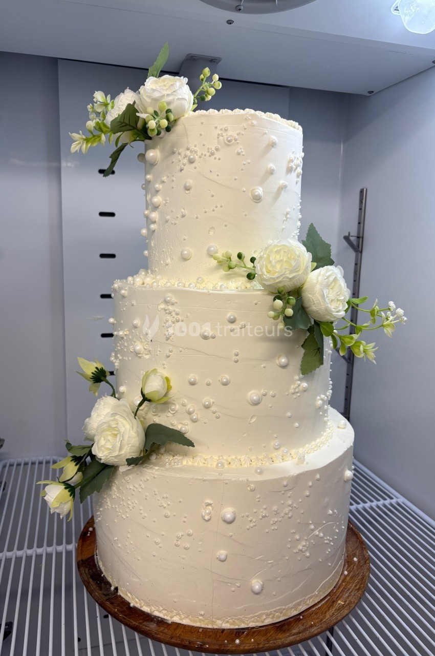Gâteau de mariage à trois étages décoré de fleurs blanches et de perles en glaçage, posé sur un plateau en bois.