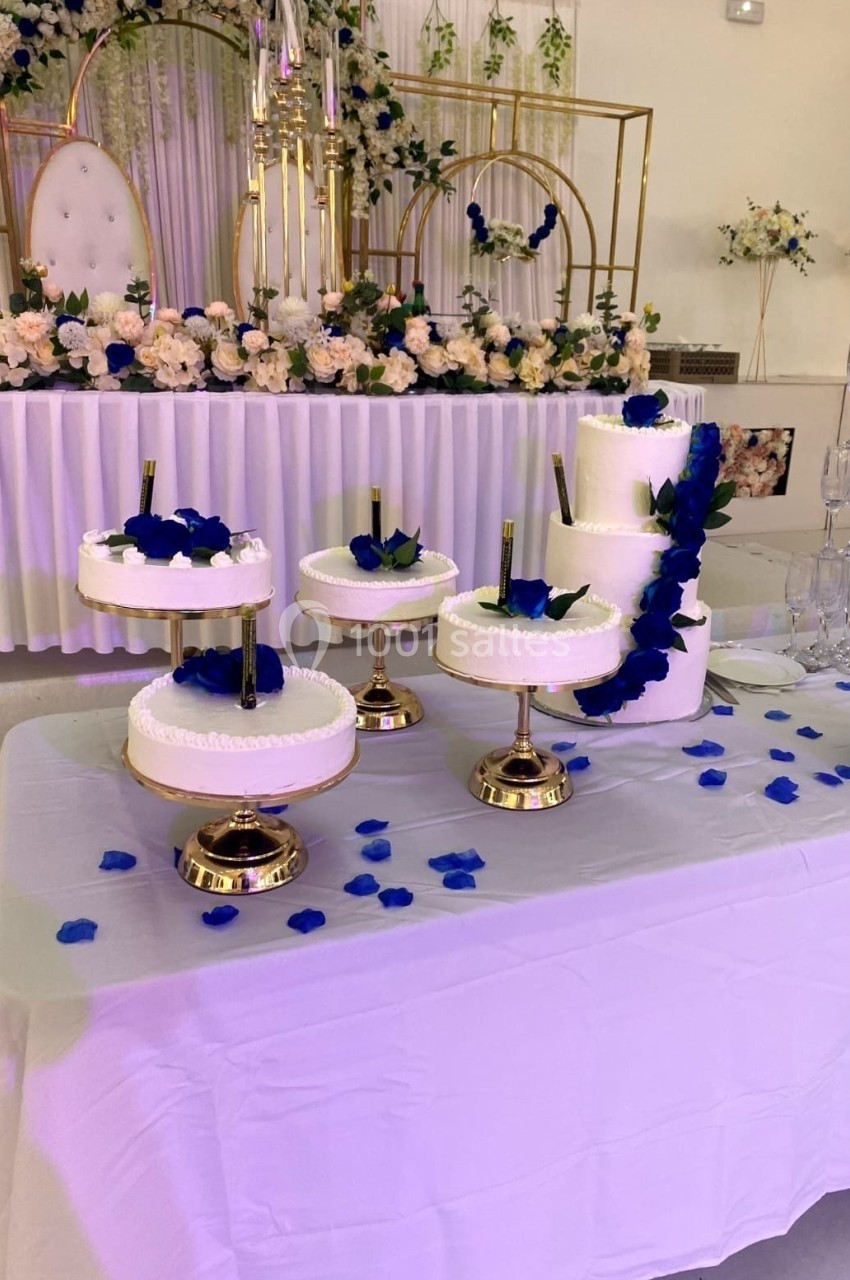 Table décorée avec plusieurs gâteaux blancs sur des présentoirs dorés, ornés de pétales et fleurs bleues.
