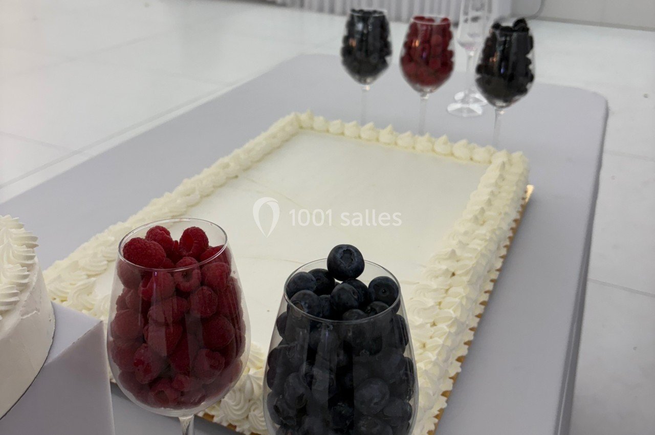 Gâteau rectangulaire décoré de crème blanche, entouré de verres contenant des framboises, myrtilles et autres fruits rouges.